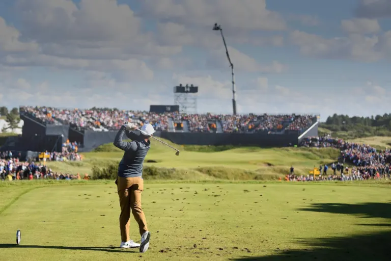 A player drives from the tee in front of a packed grandstand at The Open